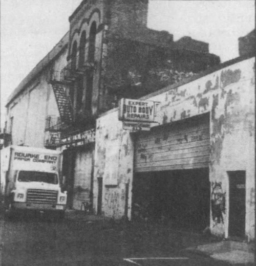 Photograph of a city block featuring a one-story white building with a garage door and a sign stating "Expert Auto Body Repairs".